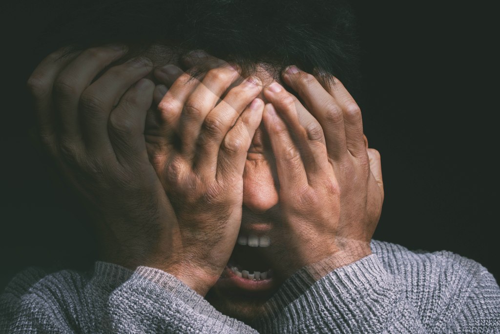 Young man in mental distress, covering his face with his hands on a black background